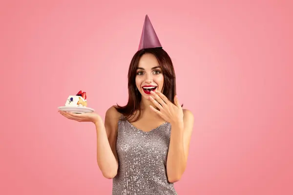 パーティーキャップでハッピーミレニアルな慎重なブルネット女性 ドレスメイク キャンドル付きケーキ ピンクの背景 スタジオで分離 ドリーム オファー お祝いの誕生日パーティー — ストック写真