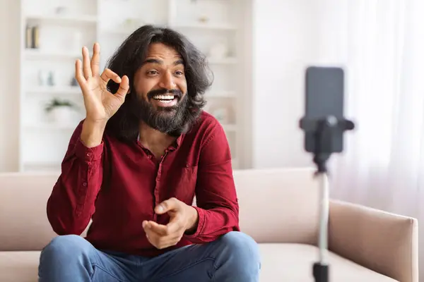 英俊英俊的长发印地安人影响着从家里流出来的人 坐在沙发上 在智能手机摄像机前摆出一副不错的姿势 著名博主用电话录影 — 图库照片