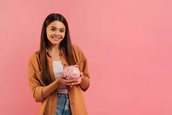 笑顔10代の少女がピンクのピギーバンクを持って 財政的責任と節約の概念を実証し 幸せな女性ティーンは ピンクのスタジオの背景にポーズカジュアルな服を着て スペースをコピー — ストック写真