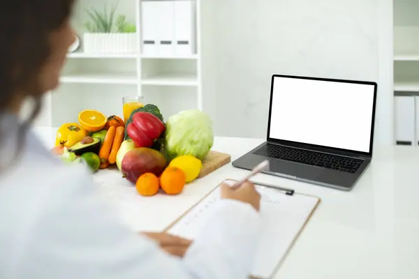 Doctor dietician have online consultation, sitting at desk with fresh organic fruits and vegetables, using computer with white screen, laptop, taking notes in clipboard, mockup, copy space