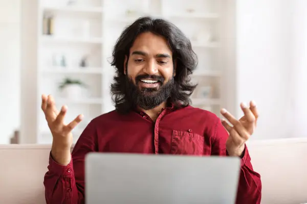 Portrait of bearded long-haired young indian man have video call on laptop. Eastern guy sitting on couch at home, looking at computer screen, smiling and gesturing. Telecommunication concept