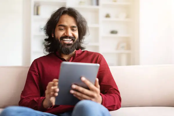 Modern gadgets and entertainment concept. Relaxed smiling bearded millennial eastern guy sitting on couch and scrolling, gaming online, using digital tablet at home, copy space