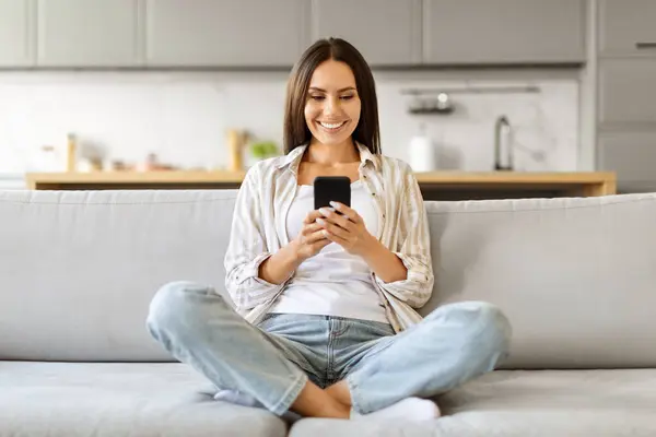 Νεαρή Χαμογελαστή Γυναίκα Smartphone Ξεκουράζεται Στον Καναπέ Στο Σπίτι Ευτυχισμένη — Φωτογραφία Αρχείου