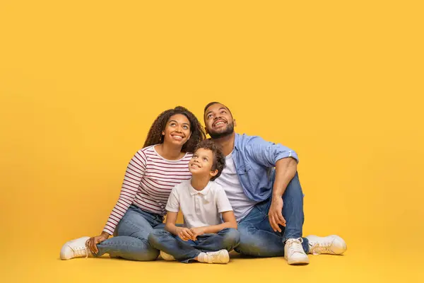 興味深いオファー 3人の笑顔のアフリカ系アメリカ人の家族は 興奮と共にコピースペースで上向きを見ています 黒人の親と彼らの息子は一緒にイエロースタジオの背景に座っています — ストック写真