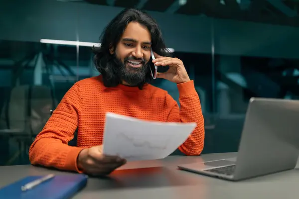 Wealthy cheerful young indian man entrepreneur working at office, sitting at table, having phone conversation, using laptop, checking papers marketing report, copy space. Business concept