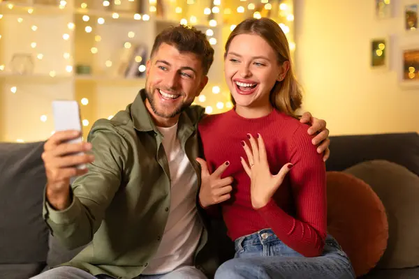エキサイティングな若い女性と笑顔の男は 彼女が結婚の提案の後に彼女の手を示すように バックグラウンドでお祝いのライト — ストック写真