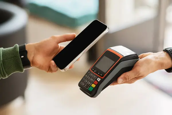 Customer using banking mobile app for payment at cafe restaurant, cashless technology and credit card payment concept. Cropped black man hand hold smartphome next to terminal, contactless payment