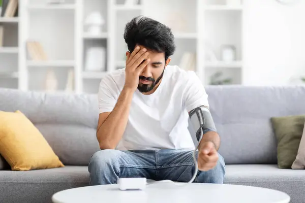 stock image Concerned indian man with blood pressure cuff on his arm monitoring his health at home, young eastern male feeling unwell, sitting on couch with distressed expression, having serious wellness issue