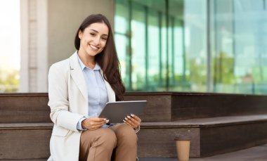 Neşeli genç profesyonel bir kadın dijital tabletle bankta oturuyor, bileğinde akıllı bir kol, kahve fincanının yanında, gülümsüyor ve arka planda modern cam bina ile kameraya bakıyor.