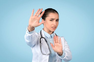 Kadın doktor jesti durduruyor, uyarıda bulunuyor veya sağlık güvenliği nedeniyle davranışları durduruyor, endişeli bir ifadeyle, mavi arka plan.