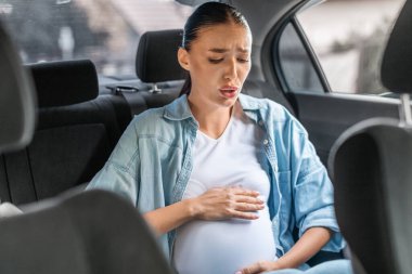 Young pregnant woman experiencing Braxton Hicks contractions sitting in back seat of car, touching belly feeling discomfort and pain. Urgency and challenges of pregnancy during transportation clipart