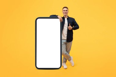 Pozitif stil sahibi, büyük akıllı telefonlu, boş ekranlı poz veren, baş parmağını kaldıran ve gülümseyen, en yeni mobil uygulamayı tavsiye eden, sarı stüdyo arka planı, model, tam boy