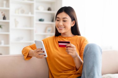 Hızlı Çevrimiçi Alışveriş. Gülümseyen Çinli kadın banka kartını tutuyor ve cep telefonu kullanıyor. Oturma odasındaki koltukta oturuyor. Akıllı telefon ekranına bakıyor.
