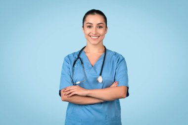 Mavi önlüklü, kollarını kavuşturmuş, steteskop takmış, kendine güvenen ve gülümseyen kadın hemşire. Sağlık sektöründe profesyonelliği temsil ediyor. Mavi arka plan.