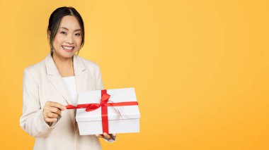 Bin yıllık takım elbiseli Koreli bayanın sarı stüdyo arka planında izole edilmiş hediye paketi kurdelesi açmasına sevindim, panorama. Tatil hediyesi, doğum günü kutlaması, kutlama yıldönümü.