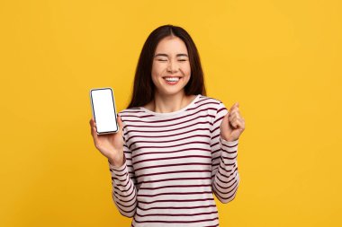 Aşırı mutlu, duygusal Asyalı bir kadın telefonu beyaz ekranlı gösteriyor, sarı duvar arkasından yüzünü ekşitiyor ve işaret ediyor, harika bir online teklif sunuyor. Nakit para