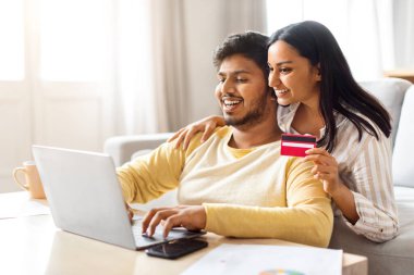 İnternet alışverişleri. Laptop ve kredi kartıyla gülümseyen Hintli çiftin portresi, mutlu genç Doğu eşleri evde dinlenirken online alışveriş için bilgisayar kullanıyorlar,