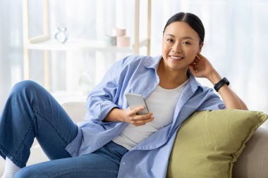Rahat mutlu milenyum Çinli kadın rahat rahat kıyafetler giyiyor elinde akıllı telefonuyla koltukta oturuyor, kameraya gülümsüyor, internetten alışveriş yapıyor, web sörfü yapıyor, uzay kopyaları yapıyor.