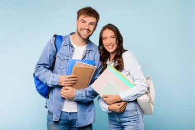Kot pantolonlu mutlu genç çift, sırt çantalı ve kitap tutan bir adam, ve her ikisi de parlak mavi arka planda, üniversitede güne hazır klasörleri tutan bir kadın.