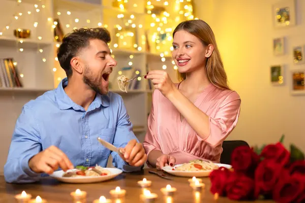 一个快乐的女人正在给她的伴侣吃意大利面 两个人都在烛光下享受着浪漫的晚餐 背景是一束玫瑰和节日的彩灯 — 图库照片