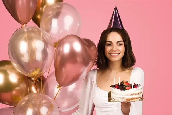 白いブラウスとパーティー帽子のラディアント若い白い白い白いケーキの女性は ピンクの背景に光沢のある風船の集団に囲まれたキャンドルで誕生日ケーキを喜んで保持しています — ストック写真