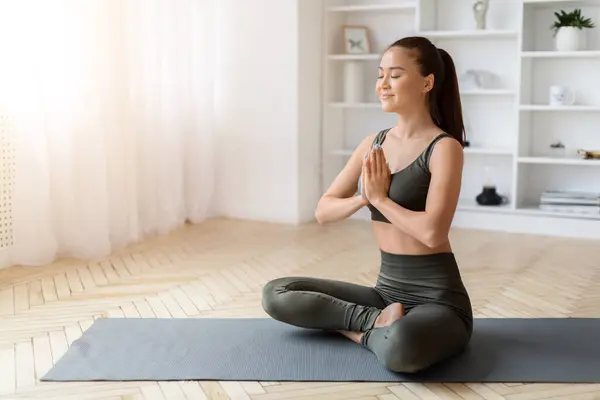 Content asian woman practicing yoga at home, sitting in namaste pose, beautiful calm korean female meditating with closed eyes on mat in her well-lit living room, embodying peace and mindfulness
