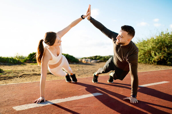 Man and woman in fitness gear sharing high-five while performing side planks on track, showcasing teamwork and encouragement in exercise