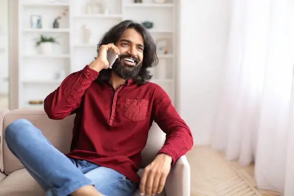 快乐的年轻英俊的 留着胡子的印度人坐在沙发上 在家里和朋友或情人通电话 分享好消息 看广告的复制空间 — 图库照片