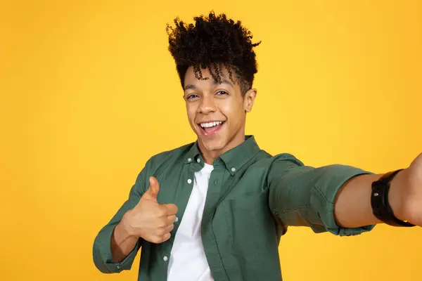 Positive handsome young black guy with stylish hairstyle wearing green shirt taking selfie on yellow background studio background, showing thumb up and smiling at camera, copy space