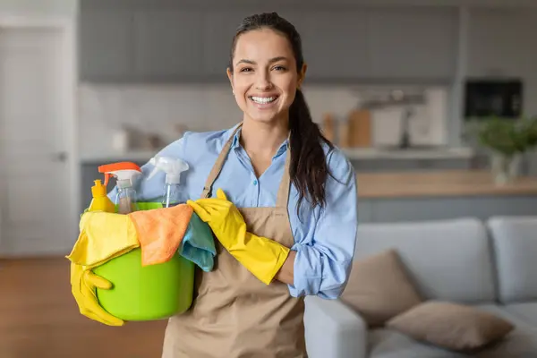 Confident young woman with beaming smile holding green bucket filled with colorful cleaning supplies and cloths, wearing yellow gloves and beige apron