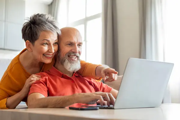 Happy senior couple websurfing on laptop together while relaxing at home, tech-savvy elderly spouses using computer in living room interior, browsing internet, shopping online, copy space