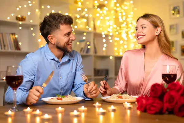 Cheerful couple sharing laugh over romantic dinner with red wine, pasta, and bouquet of roses, illuminated by warm candlelight, enjoying evening