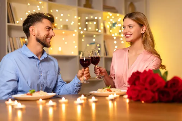 ハッピーカップル笑顔とロマンチックなキャンドルライトディナーでワイングラスを凝縮 バラの美しい花束とバックグラウンドでのお祝いのライト — ストック写真