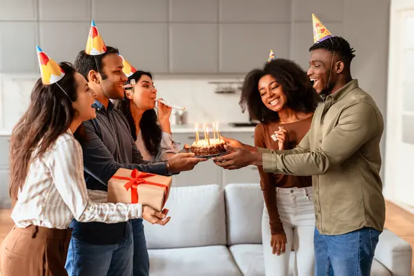 Diverse group of students celebrating birthday of a friend at home, joyfully presenting gifts and cake with burning candles in the living room interior. B-day surprise party concept