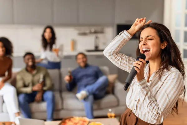 Young student lady singing in microphone during students karaoke party, friends celebrating weekend at modern apartment indoors, selective focus on woman singer. Fun and parties