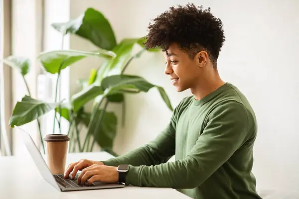 陽気な若いアフリカ系アメリカ人男性は カフェインテリアのコーヒーカップでテーブルにノートパソコンを入力します リモートワークでの独立した請負業者 職場でのデジタルノマド ブロガーとチャット サイドビュー — ストック写真
