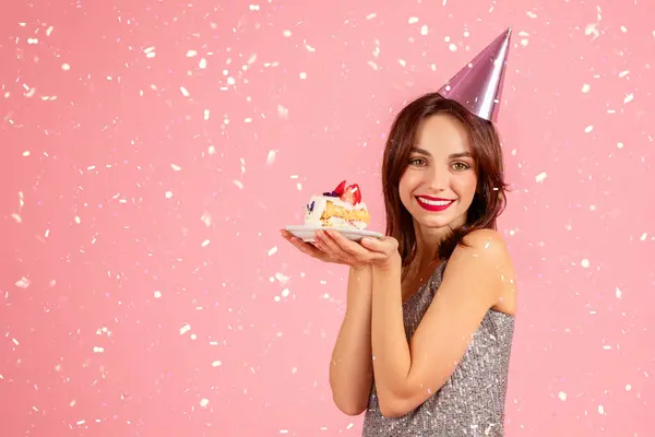 輝くドレスとパーティーの帽子で輝くミレニアル世代の白人女性は 彼女の周りに落ちる菓子と誕生日ケーキの一部を保持し ピンクの背景に対して喜んで祝います — ストック写真