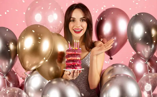 輝くドレスを着た明るい笑顔と赤い口紅を持つ女性は 金属風船と菓子のお祝いの背景の中でろうそくとドーナツのスタックを提示しています — ストック写真
