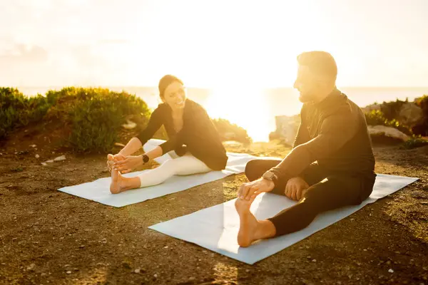 Cheerful couple enjoying their yoga routine, stretching on mats by the sea, with the golden sunlight of dusk creating warm, inviting atmosphere