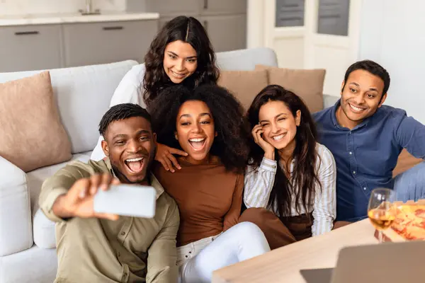 Group of young diverse friends taking selfies and making video calls on mobile phone, having fun weekend at home, gathered in modern living room. Youth and gadgets concept