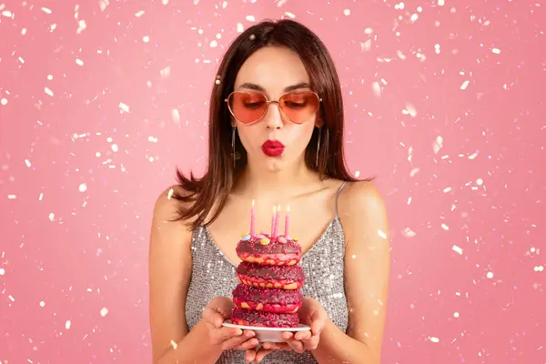 ハッピーミレニアル世代の白人女性がドレスを着て ろうそくを吹き飛ばすメガネは ピンクの背景 スタジオで隔離されたケーキドーナツで願いを作ります セレブレーションホリデー セッティと誕生日パーティー — ストック写真