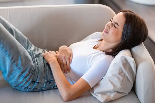 Periods and abdomen pain. Lady suffering from abdominal discomfort, having painful menstruation cramps, bad stomach condition, while lying on sofa in modern living room interior