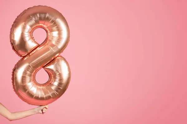 An anonymous persons hand holding a large, shiny rose gold number eight balloon on a soft pink background, possibly for a birthday or anniversary celebration