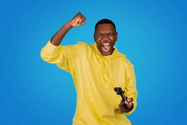 黄色いパーカーの元気な黒人男性は勝利のジェスチャーで彼の拳を切り 明るい青い背景に対してゲームコントローラーを保持しながら興奮を叫びます — ストック写真