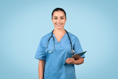 Mavi üniformalı kadın doktor, elinde bir pano tutarken kameraya gülümsüyor. Profesyonel sağlık hizmetlerini sembolize ediyor. Mavi stüdyo geçmişi.