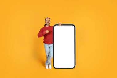 Pozitif çocuk hippi pozu boş ekranlı, telefonu gösteriyor ve gülümsüyor, en yeni mobil uygulamayı tavsiye ediyor, sarı turuncu stüdyo arka planı, model, tam boy