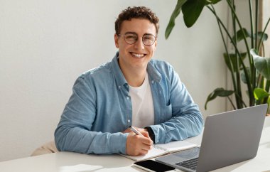 Gözlüklü neşeli bin yıllık beyaz erkek öğrenci, bilgisayar kullanıyor, çalışıyor, internette çalışıyor, kafede deftere yazıyor. Dijital göçebe, serbest çalışan, teknoloji ile fikir ve girişim yarat