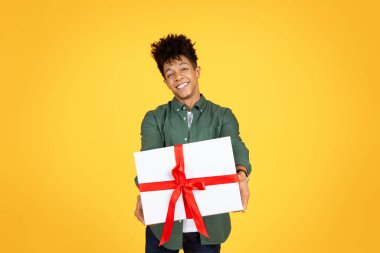 Happy handsome young black guy giving present white gift box with red bow and smiling at camera, posing isolated on yellow background. Birthday party, Valentines day celebration clipart
