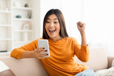 Neşeli mutlu Asyalı kadın başarıyı evinde akıllı telefonuyla kutluyor, iyi haberler alıyor, heyecanla bağırıyor ve elini kaldırıyor, boşluğu kopyalıyor.