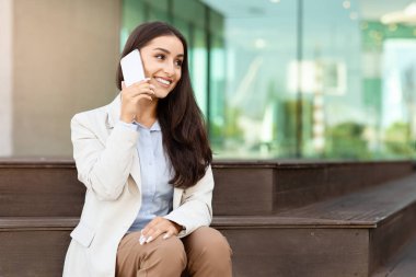 Açık hava bankında otururken akıllı bir telefonla konuşan açık renk ceketli, gülümseyen iş kadını arka planda bulanık bir modern ofis binası ön yüzü başarılı genç profesyonelleri somutlaştırıyor.